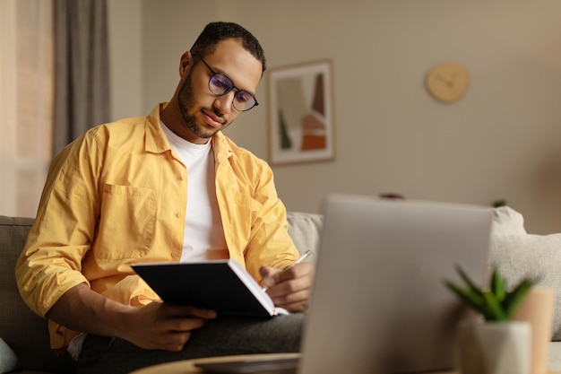 Fokussierter junger afroamerikanischer Mann mit Laptop, der online arbeitet und sich während des Geschäftstreffens Notizen macht