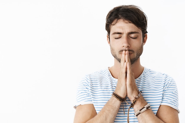 Fokussierter junger hoffnungsvoller hispanischer Kerl mit Bart in gestreiftem T-Shirt, der die Hände im Gebet über den Lippen hält und mit geschlossenen Augen betet, mit Gott spricht, Wünsche macht und auf ein Wunder wartet