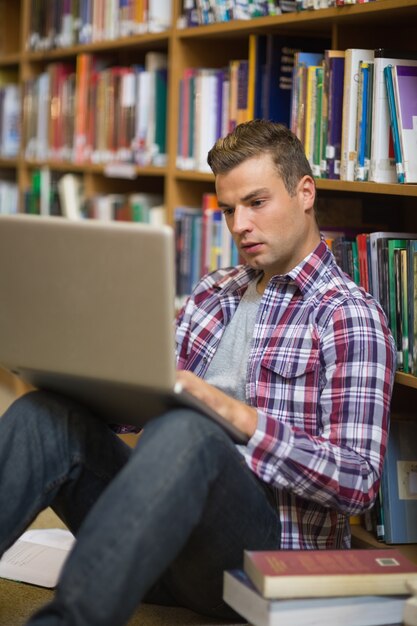 Fokussierter junger Student, der auf Bibliotheksboden unter Verwendung des Laptops sitzt