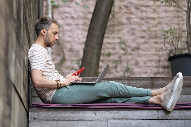 Fokussierter junger Unternehmer, der draußen mit Laptop und Smartphone sitzt, genießt den Arbeitstag unter freiem Himmel