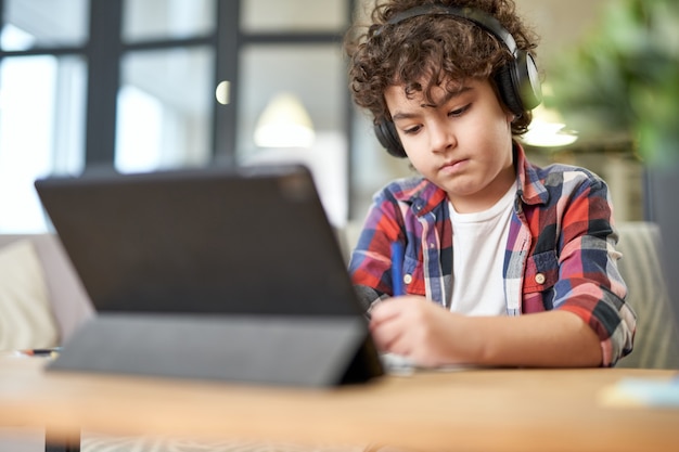 Fokussierter lateinamerikanischer Schuljunge, der Kopfhörer trägt, Notizen macht, digitale Tablette verwendet, während er zu Hause lernt. Online-Bildung, Technologie, Homeschooling