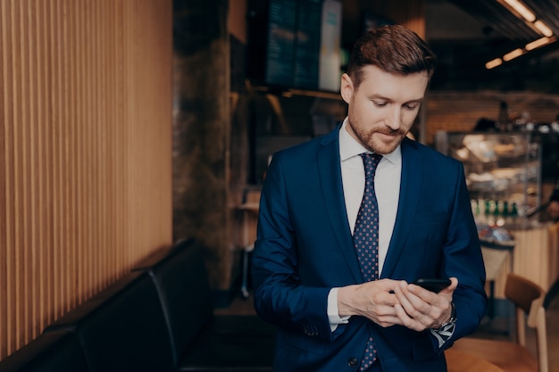 Fokussierter männlicher Investor im blauen Anzug, der über Börsenkurse informiert wird, sein Telefon überprüft, Nachrichten ansieht und entscheidet, ob er investieren soll oder nicht, allein im Café stehen