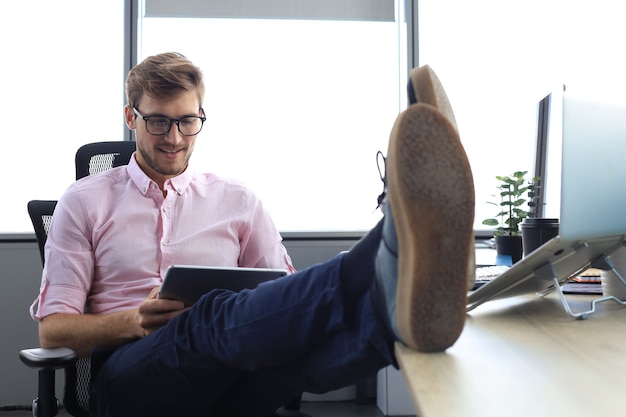 Fokussierter moderner Geschäftsmann, der in seinem modernen Büro mit digitalem Tablet arbeitet, Beine auf dem Schreibtisch.