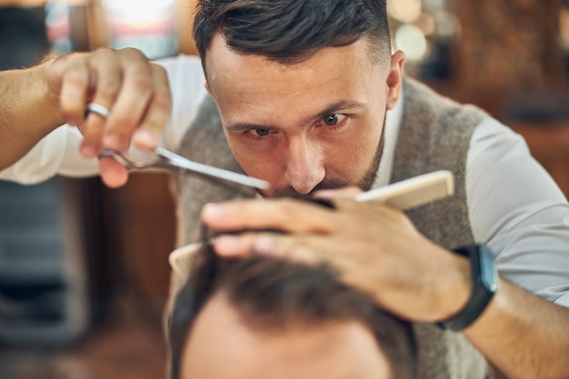 Fokussierter Profifriseur, der sich die Haare seines Besuchers genau ansieht, während er an seinem neuen Schnitt arbeitet