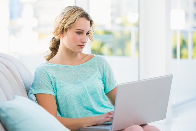 Fokussiertes blondes Sitzen auf Couch unter Verwendung des Laptops