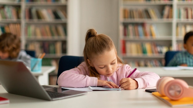 Fokussiertes kaukasisches Schülermädchen schreibt in einem Schreibbuch, während es einen Laptop benutzt und an einem Schreibtisch sitzt.