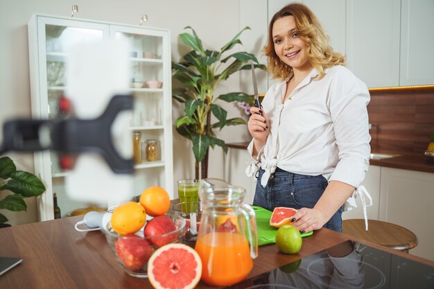 Folgen Sie mir. Fröhliches lockiges Mädchen, das Positivität ausdrückt, während es frischen Saft für ihr Mittagessen macht