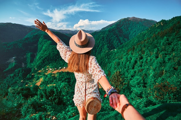 Folgen Sie mir Konzept und reisen zusammen. Mädchenreisender mit Hut, runder Strohtasche und kurzem Overall hält die Hand des Freundes und führt zu einem grünen Urlaub in den großen Bergen