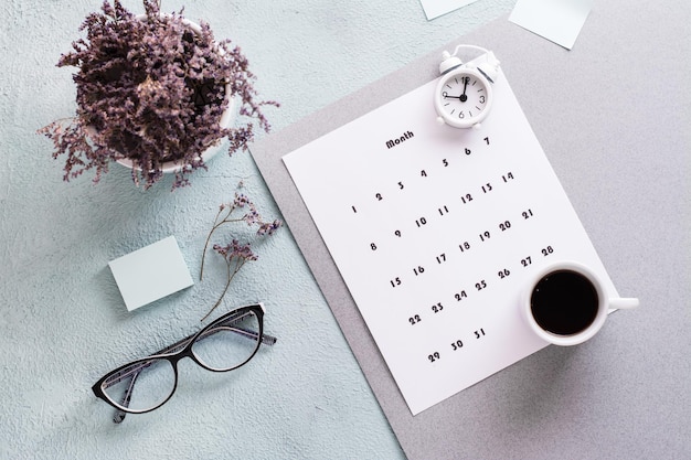 Folha de calendário mensal, xícara de café, despertador, copos e um buquê de flores na mesa. Planejamento e organização do tempo