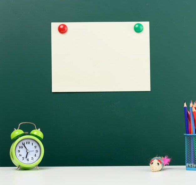 Foto folha de papel em branco afixada em um quadro de giz verde despertador e lápis multicolor de volta para a escola