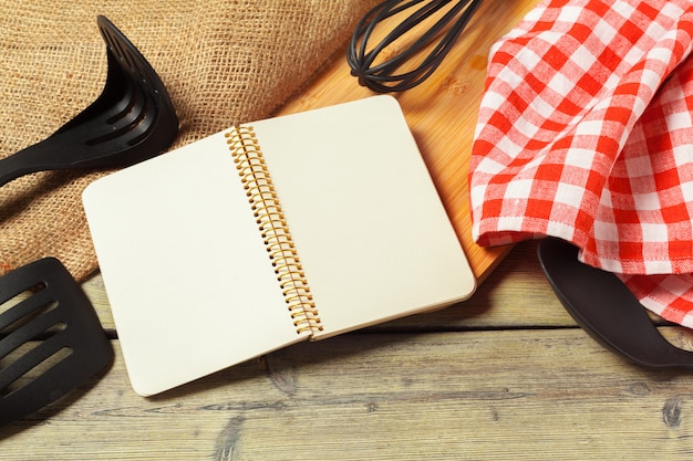 Folha em branco do bloco de notas aberto e utensílios de cozinha na mesa com toalha de mesa, cópia espaço