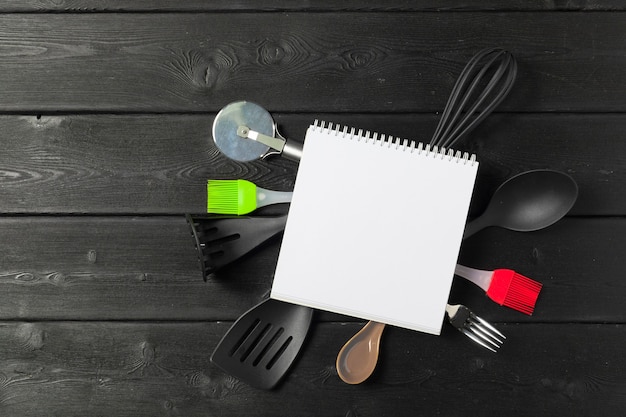 Folha em branco do bloco de notas aberto e utensílios de cozinha na mesa com toalha de mesa, espaço de cópia