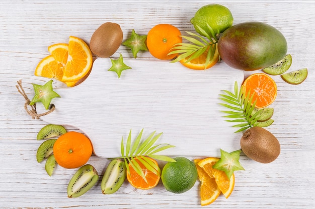 Folhas de palmeira verde e fatias de frutas tropicais frescas com uma tábua de madeira vazia na mesa branca