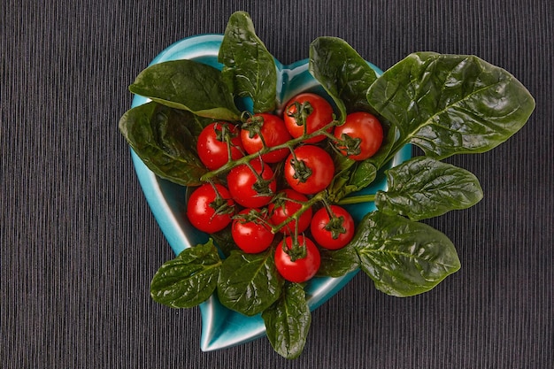 Folhas verdes de espinafre e tomate cereja vermelho em um prato azul em forma de coração em uma toalha de mesa têxtil preta