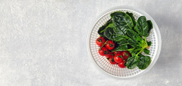 Folhas verdes lavadas de espinafre e tomate cereja em um galho em uma peneira branca de plástico sobre uma mesa de pedra clara