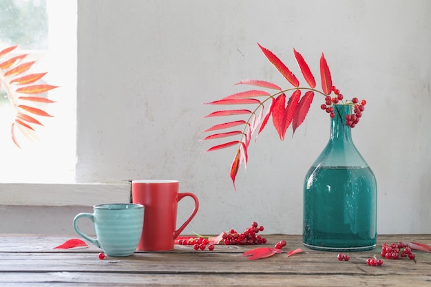 Folhas vermelhas de outono em um vaso de vidro e bagas de viburnum e uma xícara de chá na mesa de madeira