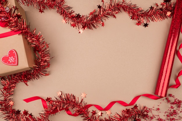 Fondo aislado Un regalo en papel artesanal, con una cinta roja sobre la cual reposan medias de dulces, mitones y un corazón de papel, está rodeado de oropel rojo, rollos con empaque.