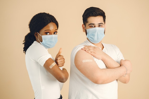 Fondo beige aislado. Mujer africana y hombre con máscaras mostrando los pulgares para arriba. Vacunación contra covid.