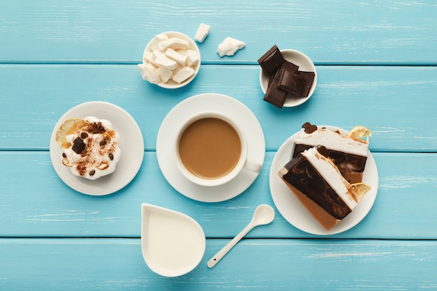 Fondo de café de la mañana. Taza de capuchino de borde, tarro de leche, chocolate, pasteles pequeños y azúcar refinada en la mesa de madera rústica azul, espacio de copia, vista superior