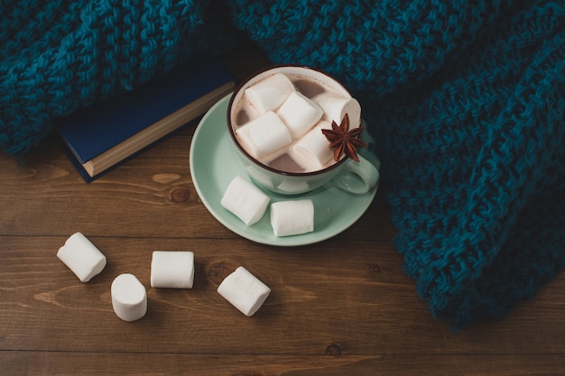 Fondo de casa de invierno - taza de chocolate caliente con malvaviscos y suéter de punto azul cálido en la mesa de madera.