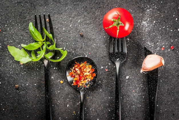 Fondo de cocina Un conjunto de artículos de mesa negros: tenedores, cuchillo y cucharas con hierbas frescas (rúcula de albahaca), tomates, ajo y especias.