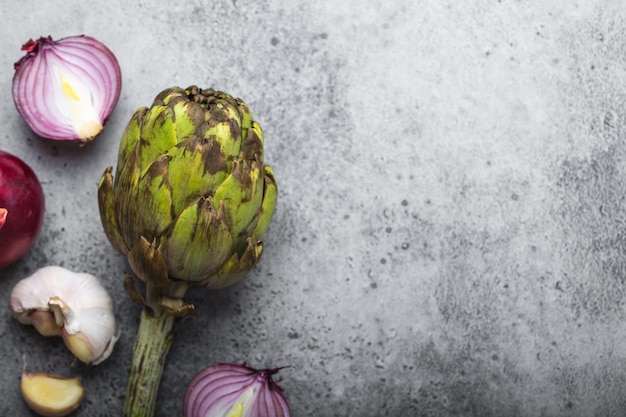 Fondo de comida con espacio libre para texto. Primer plano, vista superior de cebollas crudas cortadas, ajo y alcachofa fresca entera sobre fondo de piedra gris rústico, cocina, comida vegetariana o concepto de alimentación saludable