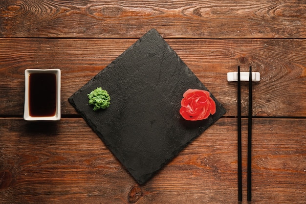 Fondo de comida japonesa. Pizarra negra vacía con jengibre y wasabi, palillos y soja servido en una mesa rústica oscura de madera, plano, vacío