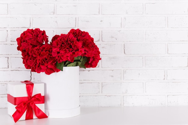 Fondo de cumpleaños o día de San Valentín - hermosas flores rojas y caja de regalo sobre pared de ladrillo blanco