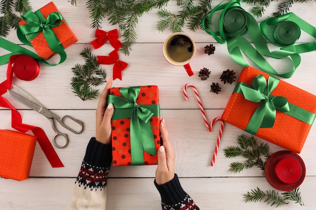 Fondo de envoltura de regalos. Las manos femeninas empaquetan el regalo de Navidad en papel punteado rojo, decoran con cinta de raso en la mesa de madera rústica desordenada blanca. Concepto de vacaciones de invierno.