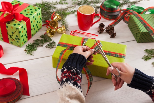 Fondo de envoltura de regalos. Las manos femeninas empaquetan el regalo de Navidad en papel verde, decoran con cinta de raso en la mesa de madera rústica desordenada blanca. Concepto de vacaciones de invierno.