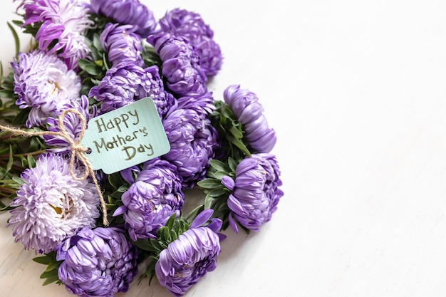 Fondo festivo con un ramo de crisantemos azules y una tarjeta de felicitación con un deseo de un feliz día de la madre sobre un fondo blanco, espacio de copia.