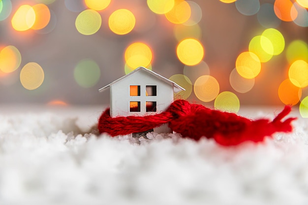 Fondo de Navidad de Adviento abstracto Casa modelo de juguete con bufanda roja de punto en la nieve con fondo de luces de guirnalda desenfocado Navidad con el concepto de familia en casa Composición de invierno de Navidad