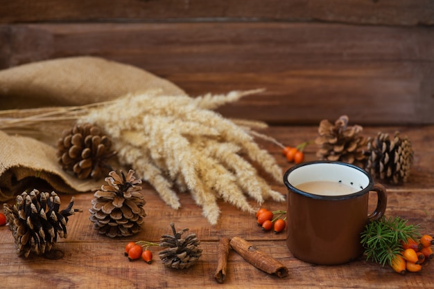 Fondo de Navidad en estilo rústico. Una taza de metal con té con leche caliente se encuentra sobre un mantel, sobre una superficie de madera entre piñas, ramas de abeto, espiguillas de trigo y rosa mosqueta. Copie el espacio, endecha plana