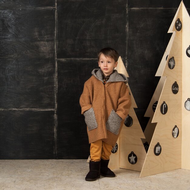 En un fondo oscuro, un niño pequeño posando con un elegante abrigo cerca de un árbol de Navidad hecho de madera