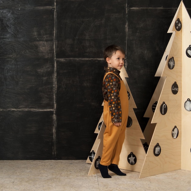 En un fondo oscuro, un niño pequeño posando con un traje de moda cerca de un árbol de Navidad hecho de madera