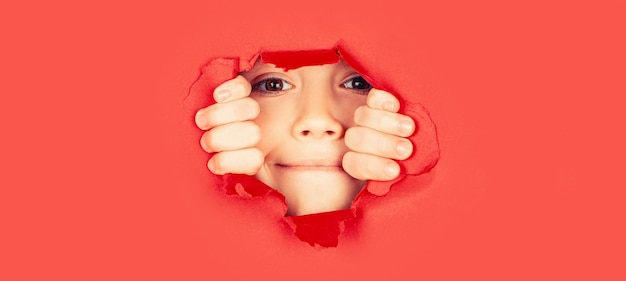 Fondo de papel roto Concepto de emociones Niño positivo con una sonrisa agradable en la cara se mantiene a través del agujero rasgado en papel rojo