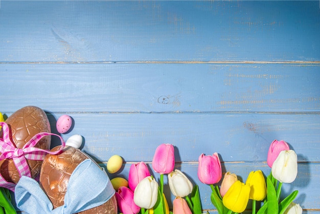 Fondo de tarjeta de felicitación de vacaciones de Pascua con huevos de conejito de Pascua de chocolate y flores de tulipán sobre fondo azul claro marco plano copia espacio vista superior