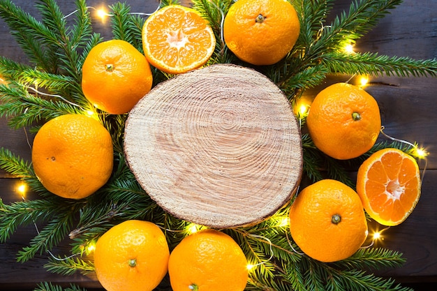 Fondo de vacaciones de año nuevo en corte redondo de árbol rodeado de mandarinas, ramas de abeto vivo y guirnaldas de luces doradas, con espacio de madera para texto. Aroma cítrico, rodajas de naranja, Navidad. marco