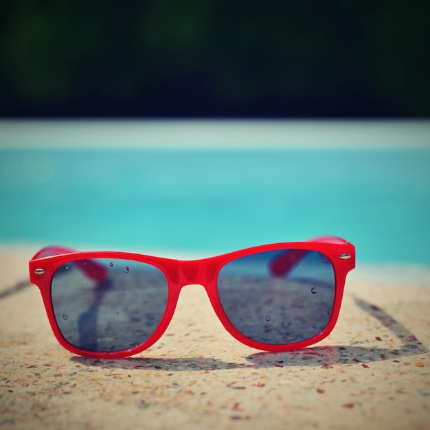 Fondo de verano Concepto para el verano y las vacaciones Gafas de sol rojas junto a la piscina en el fondo con agua azul