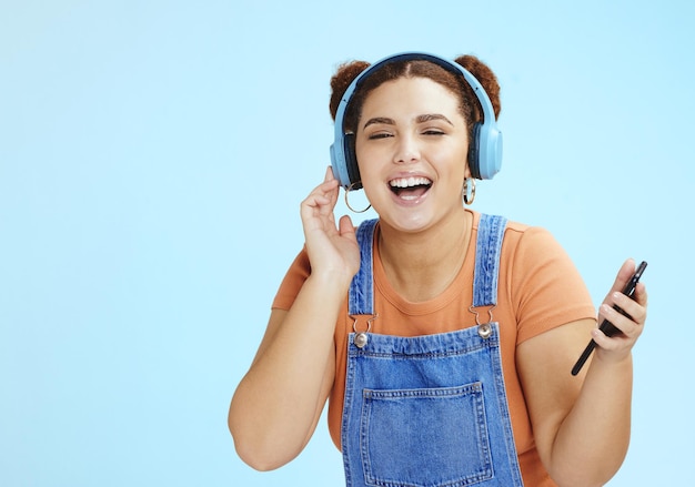 Fones de ouvido aluna e streaming de música por telefone de um modelo gen z com fundo de estúdio azul Isolado feliz e liberdade de uma pessoa ouvindo rádio na web e áudio de podcast com um sorriso