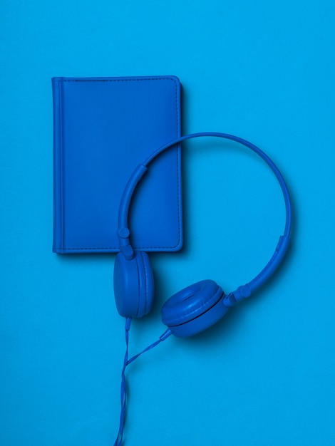 Fones de ouvido azuis com um caderno de couro azul sobre uma superfície azul. Imagem monocromática de acessórios de escritório.