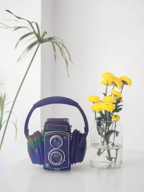 Fones de ouvido musicais em uma câmera fotográfica preta vintage colocada em uma mesa branca perto de um vaso com flores amarelas no quarto em casa