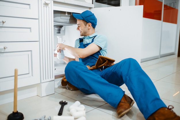 Fontanero masculino sonriente en uniforme sostiene el tubo de desagüe en la cocina. Handywoman con fregadero de reparación de bolsa de herramientas, servicio de equipamiento sanitario a domicilio