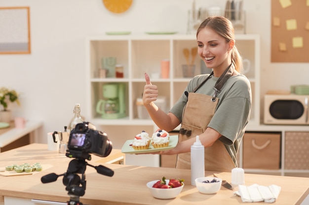 Food-Bloggerin, die gebackene Kuchen in den Händen hält und den Daumen zur Kamera zeigt, während sie das Video in der Küche dreht