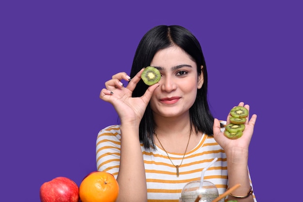 foodie chica sentada en la mesa de frutas sonriendo y esconde su ojo con kiwi modelo paquistaní indio