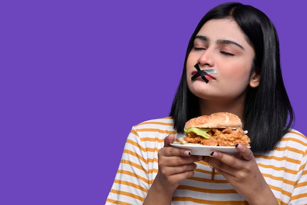 foodie girl con cinta en la boca y sosteniendo una sabrosa hamburguesa con los ojos cerrados modelo paquistaní indio