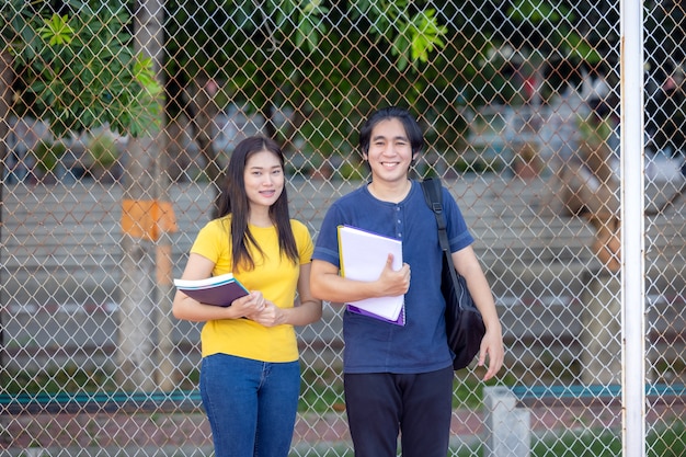 Fora da escola, um jovem casal feliz de alunos está parado ao lado de uma cerca, estudando um livro.