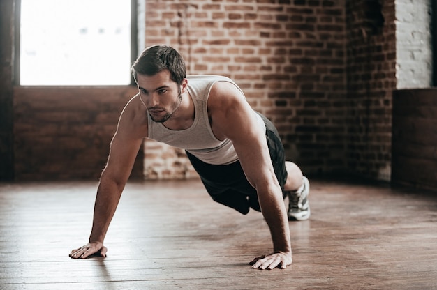 Força corporal. Homem jovem e musculoso confiante, usando roupas esportivas e fazendo flexões enquanto se exercita no chão no interior do loft