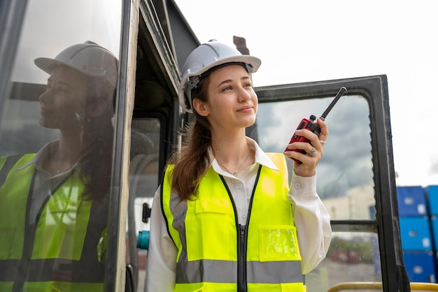 Foreman femenino permanece levantando carretilla elevadora de carga de contenedores utilizando walkie talkie para la comunicación y el control del servicio de concepto de carretilla elevadora logística de transporte Supervisor de seguridad en casco blanco