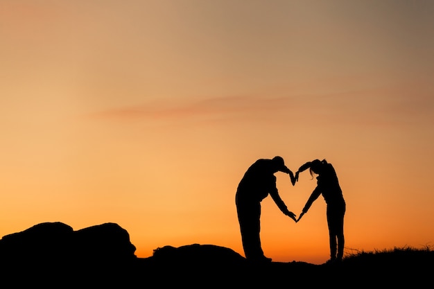 Forma de corazón conceptual, símbolo del ser humano. Silueta de mano de mujer y hombre sobre el cielo en el fondo del atardecer, metáfora del amor, día de San Valentín, romántico, pareja, boda, romance, verano o amanecer